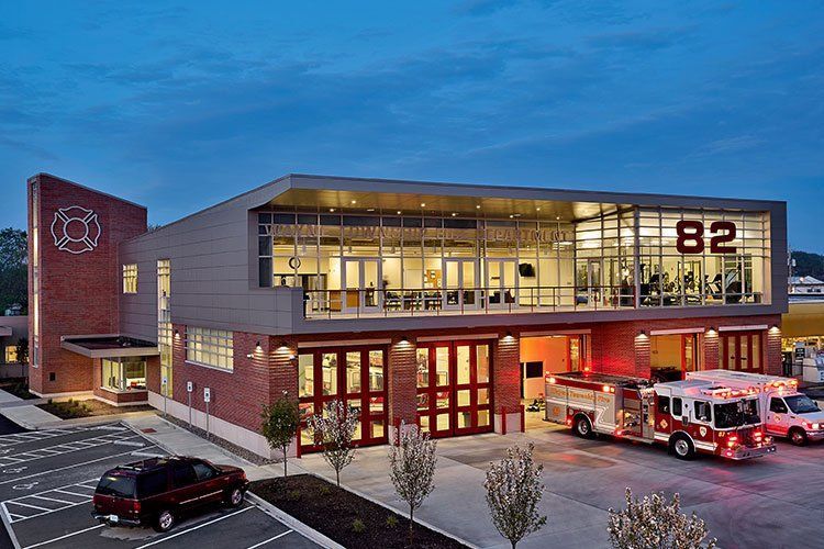 A modern brick fire station at twilight, with large glass windows, bay doors, and a fire truck parked outside.