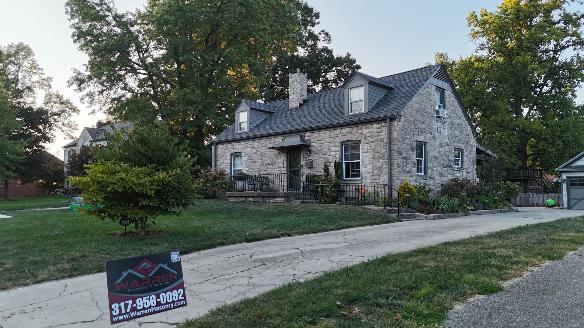 A stone house with dormer windows and a driveway, with a construction sign in the foreground, set against green trees.