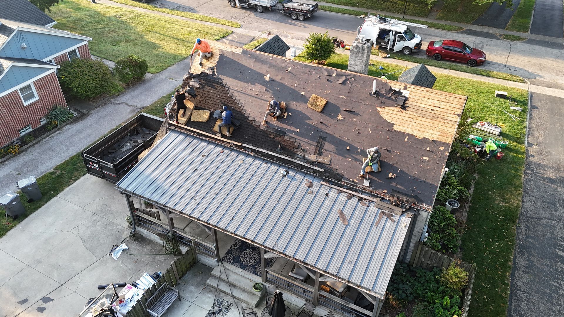 Roofers replacing shingles on a house, with a metal-roofed porch and a disposal bin in the yard.